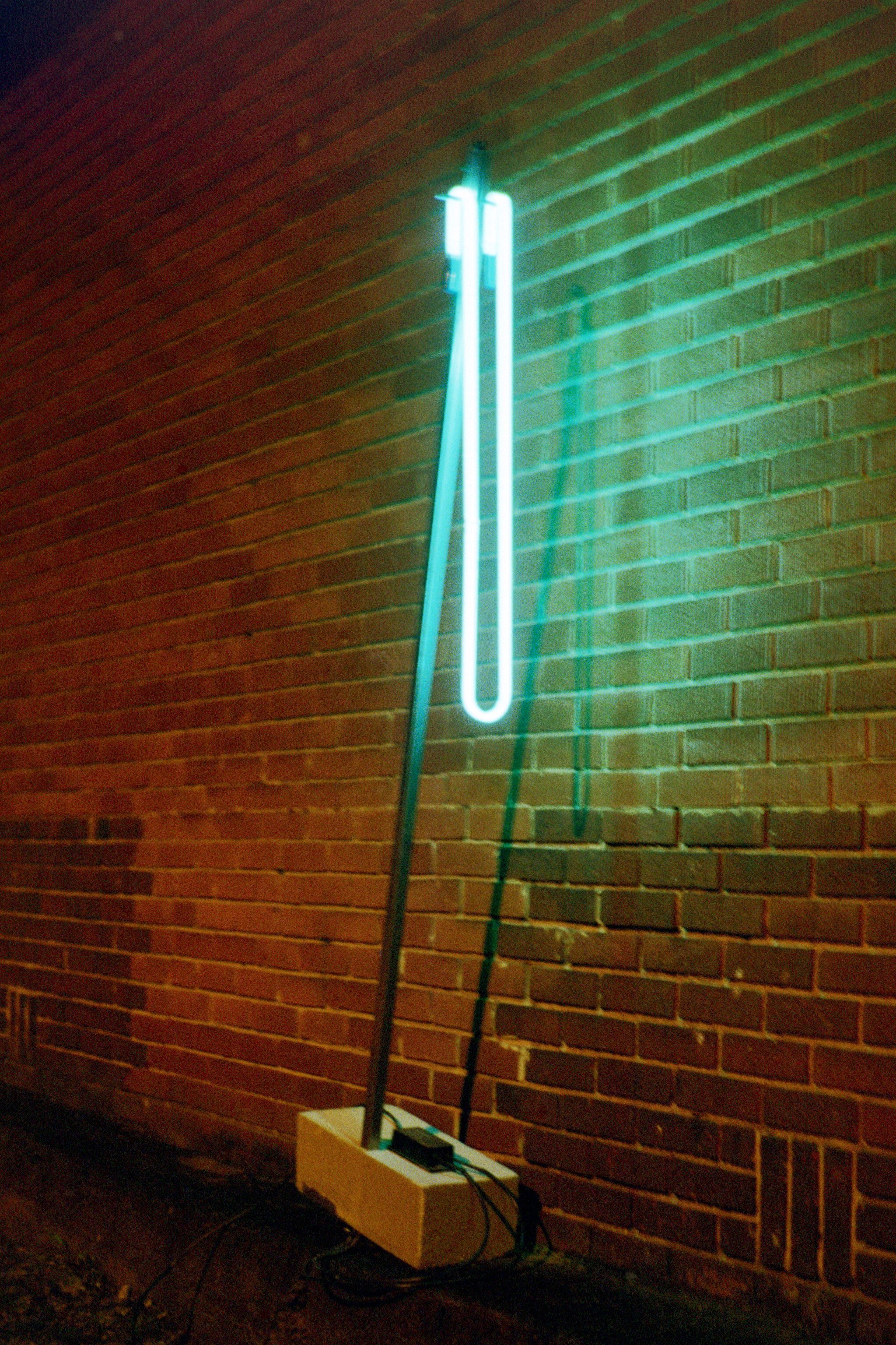 image of a glowing blue tube hanging from a tall and slender steel structure.  the steel is supported by a concrete block.  the sculpture itself sits in front of a brick wall.  The brick wall is illuminated by the blue neon as well as an orange high pressure sodium street light which is out of frame